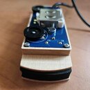 Kompaktní elektronické zařízení v dřevěném šasi s modrou základní deskou. Na horní straně jsou umístěny dva černé otočné knoflíky, jeden kovový přepí…