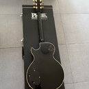 Černá elektrická kytara s bílým lemováním korpusu, položená na černém kufru. Na kufru je bílý nápis PR GUITARS a PAUL REED SMITH. Kytara má zlaté lad…