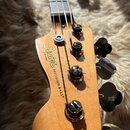 Hlava basové kytary z přírodního dřeva s nápisem Squier by Fender a označením PRECISION BASS. Jsou vidět čtyři chromované ladičky a čtyři struny vede…