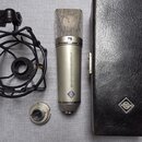 Stříbrný kondenzátorový mikrofon s kovovou síťovkou a nápisem STUDIOTECHNIK RUNDFUNK a MICROPHONE. Na těle je viditelné modré logo ve tvaru kosočtver…