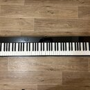 Černý digitální klavír s bílými a černými klávesami. Na levé straně je nápis CASIO a uprostřed nápis Privia. Nástroj leží na dřevěné podlaze a povrch…