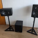Sada černých reproduktorů tvořená dvěma regálovými boxy na stojanách a jedním subwooferem. Na přední straně všech tří komponentů je viditelné logo AR…