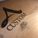 Detailní záběr na povrch mosazného činely. Je na něm černý nápis CUSTOM s velkým stylistickým písmenem A vlevo a menšími nápisy RIDE 20"/51 cm. Povrc…