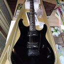 Černá elektrická kytara v kartónovém obalu. Na hmatníku je vidět bílý nápis jackson. Kytara má dva humbuckery, dvě ovládací knoflíky a jeden přepínač…