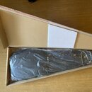 V otevřené cardboardové krabici se nachází černý nástroj nebo příslušenství zabalené v průhledné plastové fólii. V horní části krabice je umístěn bíl…