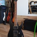 Černá elektrická kytara s dvojstupňovými humbuckery a dvěma ovládacími knoflíky. Na hlavě hрифu je viditelné logo Ibanez. V pozadí jsou v držáku umís…
