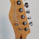 Fotografie zachycuje hlavu kytary vyrobenou ze světlého dřeva s výraznou strukturou. Na zadní straně je viditelný nápis CUSTOM GUITARS a pod ním podp…