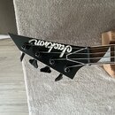 Černá hlava basové kytary s bílým nápisem Jackson. Na hlavě jsou čtyři černé ladičky. Nástroj leží na korkovém podložkovém bloku a šedém koberci.