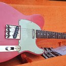 Růžová elektrická kytara typu Telecaster s bílým pickguardem, položená v otevřeném kufru s oranžovou výplní. Na hlavě hředu je viditelné logo Fender.…