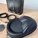 Na fotografii je černý tvrdý přepravní pouzdro na sluchátka s zipem, kable a krabice. Na krabici je nápis CROSSFADE LP2 a logo v-moda. V plastovém sá…