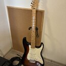 Elektrická kytara s tłem v barvě sunburst a bílým pickguardem. Na hlavě hředu je viditelné logo Fender. Kytara stojí na černém stojanu před korkovou…