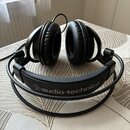 Černé náušlupky s polovrním náhlavním pásem. Na náhlavnímu pásu je bílý nápis audio-technica s doprovodným logem. Povrch polštářů a pásu vykazuje mír…