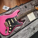 Růžová elektrická kytara v černém kufru s šedým polstrováním. Na hlavě hředu je viditelné logo Fender, na těle kytary je černý pickguard a tři snímač…