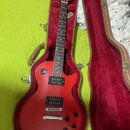 Červená elektrická kytara s tvarem single-cut v hnědém tvrdém kufru s červeným vnitřkem. Na hlavě je bílý nápis "Epiphone". Kytara má dva humbuckery,…