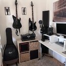 Fotografie zachicuje kout s hudebním vybavením. Na stěně visí černá elektrická kytara a černá basová kytara, na stole stojí černý kombo zesilovač s n…