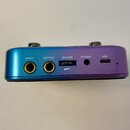 Zadní strana malého audio rozhraní s kovovým povrchem v přechodové barvě od modré po fialovou. Jsou zde viditelné konektory s nápisy INPUT, OUTPUT, M…