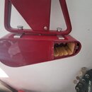Červený kovový držák na nástroje s otevřeným úložným prostorem, ve kterém jsou vidět čtyři dřevěné kolíky. V pravé dolní části je vidět hlava basové…