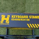 Černý papírový karton s žlutými nápisy a grafickými prvky. Je na něm vidět žluté logo ve tvaru písmene H a text KEYBOARD STAND KS400B/KS410B HERCULES…