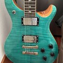Elektrická kytara s tělem z modrozelého plamenového javoru a hnědým dřevěným bokem. Nástroj má dva humbuckery, čtyři černé ovládací knoflíky a jednu…