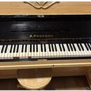 Světle hnědé dřevěné piano s černým povrchem v oblasti klávesnice. Nad klávesami je zlatý nápis A.PROKSCH a v horní části korpusu je ozdobný vyřezáva…
