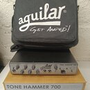 Černý basový zesilovač s nápisem aguilar TONE HAMMER 700. Přední panel obsahuje řadu ovládacích knoflíků a vstupů, včetně označení GAIN, MID LEVEL, B…