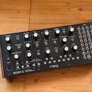 Analogový syntetizátor v dřevěném šasi s kovovým předním panelem. Na spodní hraně je nápis MOTHER-32 SEMI-MODULAR ANALOG SYNTHESIZER a logo moog. Pan…