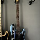 Modrá basová kytara s černým pickguardem a hnědým hmatníkem. Na hlavě nástroje je nápis Fender Precision Bass. Kytara má černý popruh s bílým nápisem…