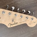 Hlava elektrické kytary z přírodního světlého dřeva s chromovanými ladičkami. Na povrchu je černý nápis Fender, STRATOCASTER a menší text WITH SYNCHR…