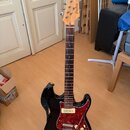 Elektrická kytara s černým bodies a červeným pickguardem v textuře želvy. Na hlavě hředu je viditelné logo Fender a nápis Stratocaster. Hmatník je tm…