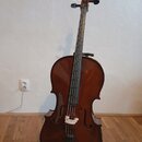Hnědý smyčkový nástroj typu violoncello s dřevěným povrchem. Nástroj je umístěn na černém stojanu před bílou stěnou. Na fotce nejsou viditelné žádné…