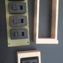 Na fotografii jsou čtyři černé elektrické spínače. Tři z nich jsou rozmístěny na zeleném obdélníkovém podkladu a jeden je umístěn v dřevěném rámečku.…