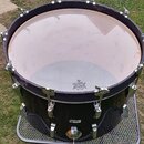 Černý buben s bílým blánkem, umístěný na kovovém mřížkovém stojanu. Na blánku je viditelný nápis REMO WEATHERKING AMBASSADOR MASS MADE IN U.S.A. Bube…