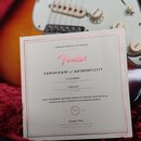 Fotografie zachicuje část elektrické kytary s bílým pickguardem a kontrolními knoflíky, na které je položen papírový certifikát. Na dokumentu je červ…