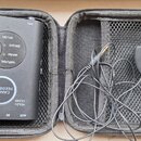 Černý přenosný efektový pedál v šedém zipovém pouzdře. Na zařízení jsou nápisy TONE, FEEDBACK CANCEL a přepínač s označením NATURAL, WARM, BRIGHT, NY…