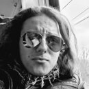 Na černobílé fotografii je zobrazen muž s dlouhými vlasy, který má na sobě koženou bundu a sluneční brýle typu aviator. V pozadí je vidět interiér vo…