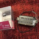Na červeném koberci leží elektrický snímač typu humbucker s kovovým krytem a připojenými vodiči. Na spodní straně snímače je nápis Made in America a…