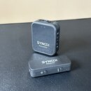 Sada bezdrátového mikrofonu tvořená černým transmitterem a receiverem. Na transmitteru je bílý nápis SYNCO Transmitter a na receiveru nápis SYNCO Rec…