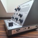 Stříbrný elektronický nástroj s nápisem Stylophone THEREMIN. Přístroj má několik černých otočných oválových knoflíků s oranžovými detaily a popisky D…