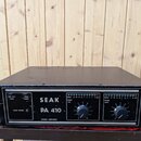 Černý výkonový zesilovač v kovovém šasi s úchopy po stranách. Na předním panelu je bílý nápis SEAK PA 410 a POWER AMPLIFIER. Jsou zde dva ovratidla V…