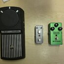 Na světlém dřevěném povrchu leží tři kytarové efekty. Zleva je zelený pedál s nápisy MXR a GT-100, vybavený dvěma otočnými knoflíky označenými jako G…