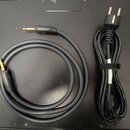 Na fotografii jsou dva černé kabely položené na tmavém povrchu. Jeden je audio kabel s dvěma zlatými 6,3mm jack konektory, na kterých je nečitelný ná…