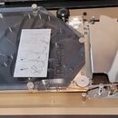 V otevřeném kufru je vidět mechanismus s kovovými a plastovými prvky. Na průhledném plastovém krytu je vyražen nápis Roland a položen je na něm papír…