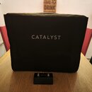Černý textilní kryt na hudební zesilovač s bílým nápisem CATALYST uprostřed. Před krytem leží černý stupňovací spínací pedál s nápisy CATALYST, CHANN…