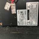 Detail zadní strany černého elektronického zařízení s dvěma průhlednými svorkami pro připojení kabelů. Na bílém štítku je uveden nápis Genius MULTIME…