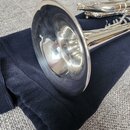 Stříbrná truba položená na tmavě modré látkové podložce. Na fotce je vidět zvon a část ventilového mechanismu s potrubím. Povrch nástroje je lesklý,…