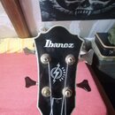 Fotografie zachycuje hlavu basové kytary v černé barvě s bílým lemováním. V horní části je bílé logo Ibanez a níže se nachází bílý symbol blesku s ná…