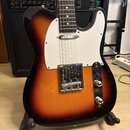 Elektrická kytara typu Telecaster s barevným přechodem od žluté přes oranžovou k černé (sunburst). Má bílý pickguard, chromovaný most s intonací a dv…