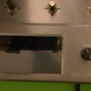 Detail stříbrného elektronického zařízení s kovovým povrchem. V horní části jsou čtyři přepínače, uprostřed je umístěn obdélníkový displej a po jeho…