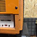 Detail zadní strany oranžového kytarového zesilovače s texturovaným povrchem. Na bílém štítku je nápis WARNING-ATTENTION a další varovné texty v angl…