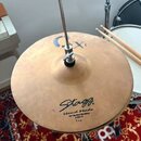Zlatý činely typu hi-hat umístěný na stojanu. Na povrchu je černý nápis Stagg, Hand Made, 13" Hi-Hat Medium, CXH-13 a Top. V horní části je viditelné…